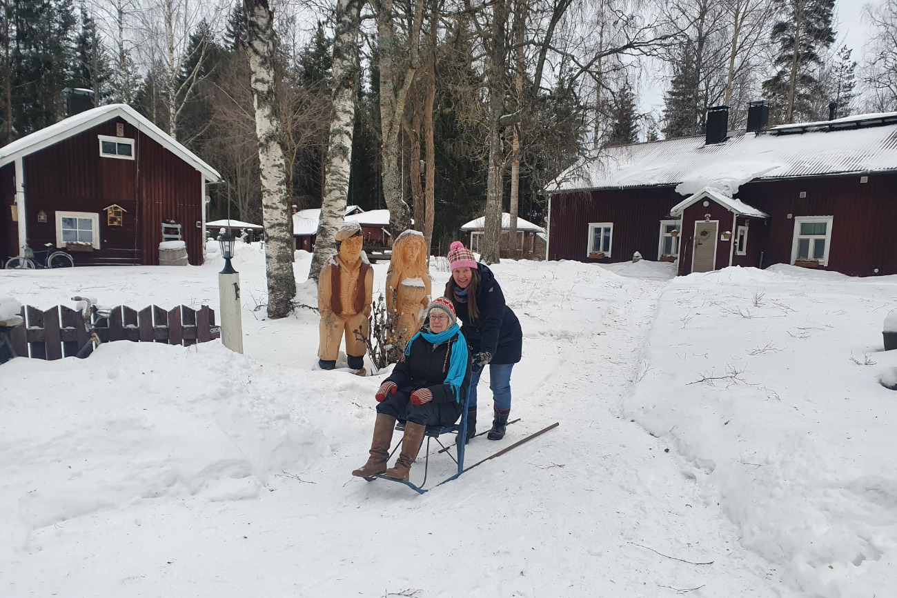 woman pushing older woman in a sled chair in the snowy forest with houses in background